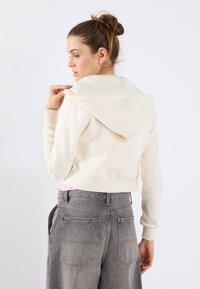 Sudadera cortada de color crema con una textura suave y una capucha grande, llevada sobre jeans de tiro alto grises con costuras visibles en los bolsillos.