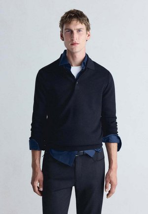 Jeune homme aux cheveux courts portant une chemise bleu marine foncé sous un pull noir et un pantalon noir, debout devant un fond uni.