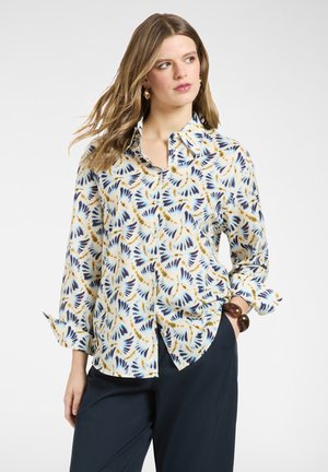 Donna che indossa una camicia bianca con bottoni decorata con motivi piumati blu e gialli, abbinata a pantaloni scuri e un braccialetto marrone.