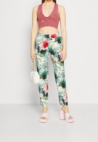 Femme portant un top court rose, un pantalon à imprimé tropical, tenant un sac à main blanc et des sandales à plateforme roses, se tenant contre un fond blanc.