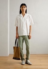 White short-sleeve shirt with a pocket, light green pants, gray sneakers, and a two-tone brown tote bag. Neutral wall and flooring background.