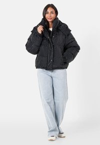 Kvinna som bär en svart vadderad jacka med horisontella ränder, ljusblå vida jeans och vita sneakers, poserar med ena handen justerande kragen.