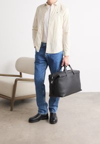Borsa in pelle nera con chiusura a zip e manici doppi, superficie testurizzata, tenuta da una persona in camicia chiara e jeans di denim.