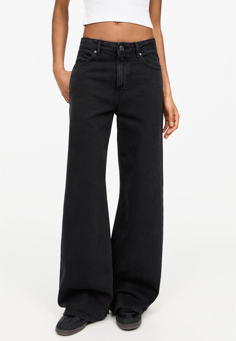 Personne portant un jean noir à jambes larges, des baskets noires et un haut blanc court, debout devant un fond uni.