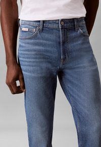 Man draagt lichtblauwe denim jeans en een wit overhemd, staand tegen een neutrale grijze achtergrond, hand ontspannen langs het lichaam.