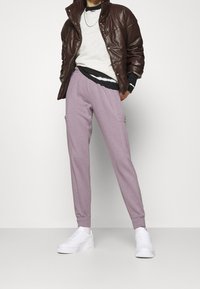 Pantaloni della tuta viola chiaro con tasche laterali, vita elastica, abbinati a un maglione grigio a girocollo, una giacca scura e sneaker bianche.