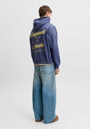Personne portant un sweat à capuche bleu marine avec un texte jaune "Originals Studio" dans le dos, un jean large bleu clair et des chaussures marron, debout de dos.