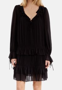 Vestido negro con volantes hecho de una tela ligera. Presenta un escote en V profundo, mangas largas con lazos y un diseño en capas con pliegues horizontales.