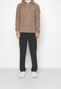 Brauner Strickpullover mit hohem Kragen und Viertel-Reißverschluss, versehen mit einem strukturierten Muster. Kombiniert mit eng anliegenden schwarzen Hosen und weißen Turnschuhen.