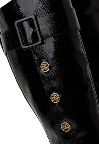 Fekete patentes bőrcsizma, térd fölé érő, széles pánttal, négyzet alakú csattal és három kör alakú arany díszítéssel az oldalon.