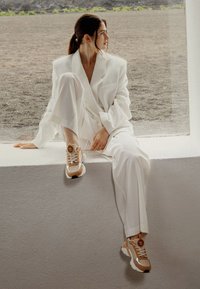 Blazer blanc surdimensionné avec un pantalon large assorti, associé à des baskets beige et blanches avec un logo et des accents texturés.