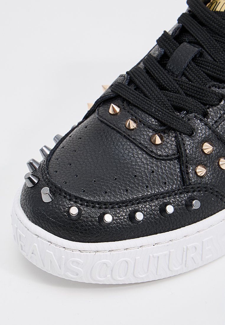 Zapatilla negra de cuero con tachuelas metálicas, superficie texturizada y suela blanca texturizada con "JEANS COUTURE" embellecida en el lateral.