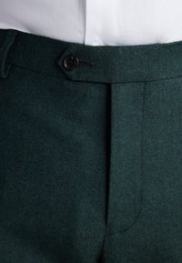 Pantaloni di lana verde scuro con linguetta in vita abbottonata sopra una camicia bianca infilata, mostrando un primo piano della zona frontale della vita.