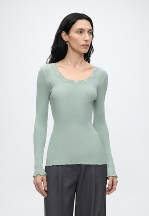 Femme aux longs cheveux foncés portant un haut côtelé vert clair à manches longues avec un col en dentelle et un pantalon large gris foncé, debout sur fond blanc.