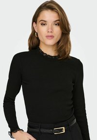 Svart langermet ribbet topp med volangdetaljer på halsen og mansjettene, med en tettsittende passform og en glatt tekstur. Brukt med et svart belte.