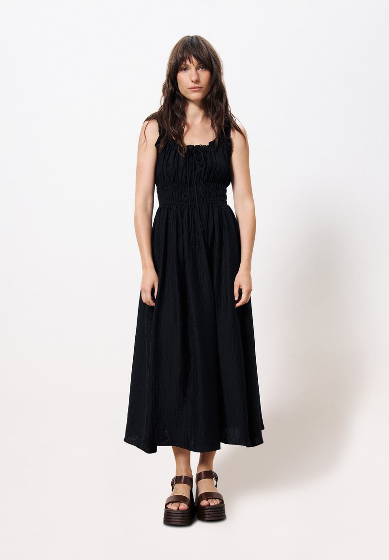 Schwarzes Maxikleid mit gerafftem Oberteil und verstellbarem Bindeband am Rundhalsausschnitt, das aus einem fließenden, strukturierten Stoff besteht und einen weiten, knöchellangen Saum hat. Braune chunky Sandalen.
