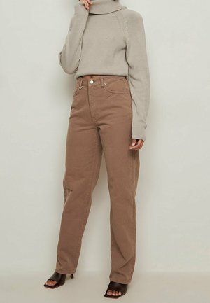 Femme portant un pull à col roulé beige, un jean marron taille haute et des sandales à talons ouvertes noires, debout devant un fond uni.