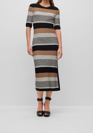 Frau trägt ein knielanges, kurzärmeliges Kleid mit horizontalen schwarzen, braunen, weißen und beigen strukturierten Streifen und schwarzen Pumps mit Knöchelriemen.