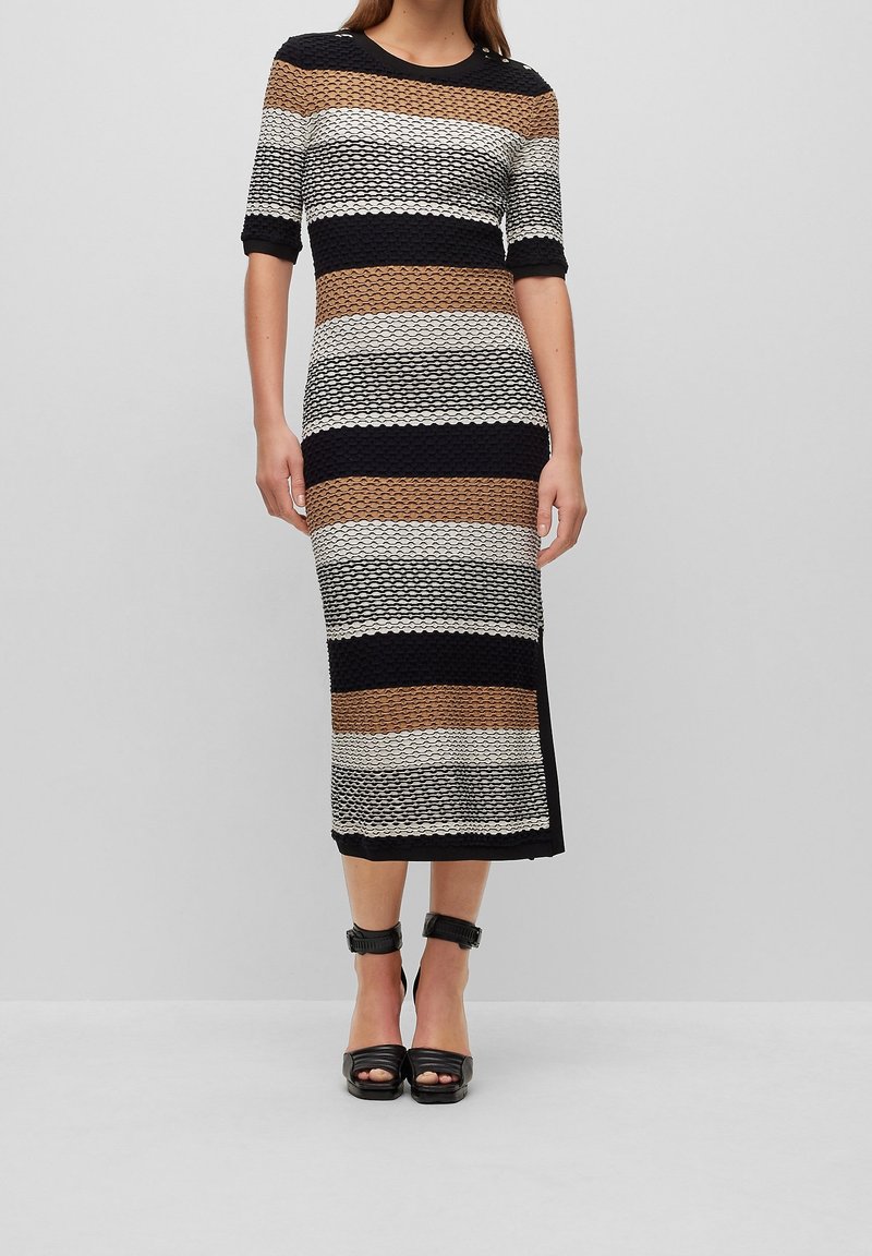 Femme portant une robe mi-longue à manches courtes avec des rayures horizontales texturées noires, marron, blanches et beiges et des talons noirs à bride à la cheville.