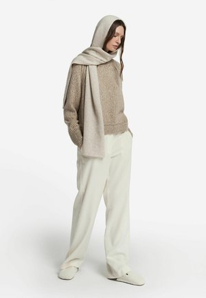 Beige strikket sweater med ribbet manchetter og kant, kombineret med hvide plisserede bukser. Modellen har et matchende beige tørklæde og bløde tøfler på.