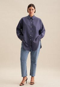 Camicetta a righe blu mare con colletto classico, maniche lunghe e spacchi laterali, abbinata a jeans dritti blu chiaro e tacchi neri.