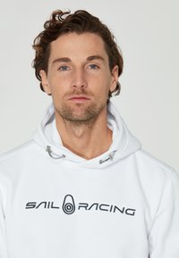 Sudadera blanca con capucha, que presenta un bolsillo delantero, cordones y el logotipo bordado "SAIL RACING" en negro. Textura suave y corte casual.