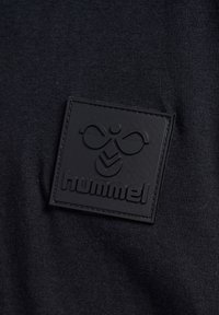 Sort tekstil med en struktureret sort læderlap, der har "hummel" mærkets logo og et stiliseret emblem, syet omkring kanterne.