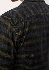 Camisa a cuadros en negro y verde oscuro con una tela texturizada, que presenta un collar puntiagudo y costuras visibles a lo largo de las uniones.