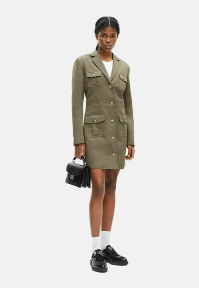 Olivgrünes, tailliertes Blazerkleid mit zwei Brusttaschen, goldenen Knöpfen und gerader Silhouette, kombiniert mit schwarzer Handtasche und Schuhen.