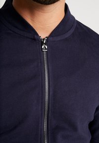 Zbliżenie osoby w ciemnoniebieskiej kurtce zapinanej na zamek z srebrnym zamkiem błyskawicznym i zaokrąglonym kołnierzem.