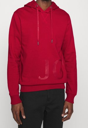Hombre con sudadera roja con capucha con cordones y letras sutilmente en relieve en el dobladillo, combinada con pantalones negros.