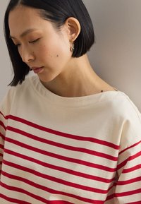 Ženska s kratkimi črnimi lasmi gleda navzdol, nosi zlate obroče za ušesa in belo majico z rdečimi vodoravnimi črtami.