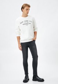 Sudadera blanca con letras negras que dicen "New York City, Brooklyn, USA", combinada con jeans oscuros de corte slim y botas negras de tobillo.