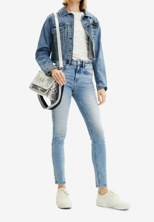 Giacca di jeans con dettaglio a stella, camicia bianca aderente, skinny jeans azzurri e sneakers bianche. Design strutturato e casual con texture a contrasto.