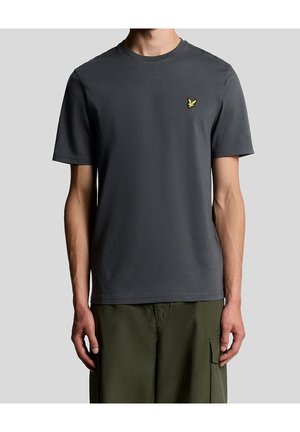 Mand iført en mørkegrå t-shirt med korte ærmer med et lille gult fuglelogo på venstre bryst og olivengrønne bukser.