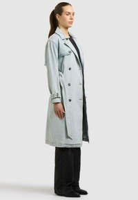 Donna in piedi di lato che indossa un trench in denim doppiopetto azzurro, pantaloni neri e scarpe nere su uno sfondo neutro.