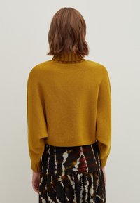 Un pull en maille jaune avec un col haut côtelé et des épaules tombantes, associé à une jupe à motif tie-dye dans des tons sombres.