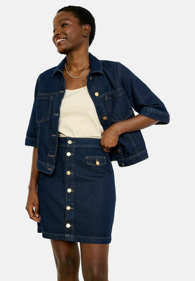 Nasmejana ženska prilagaja temno modro jeans jakno preko kremastega topa, kombinirano z usklajeno jeans krilo na gumbe, na enobarvnem ozadju.