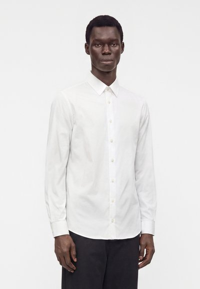 Uomo in piedi che indossa una camicia bianca a maniche lunghe con bottoni e pantaloni neri su uno sfondo bianco semplice.