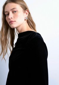 Jeune femme aux longs cheveux blonds portant un haut en velours noir et de grandes boucles d'oreilles argentées sur fond blanc.