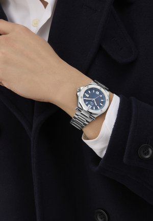 Strieborné hodinky s modrým ciferníkom a bielymi hodinovými indexmi, nerezový kovový náramok a osmihranný tvar puzdra. Nosené na ruke.