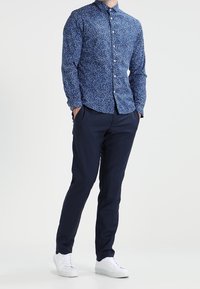 Camicia blu a fantasia con maniche lunghe, chiusura con bottoni e colletto, abbinata a pantaloni slim-fit blu scuro e sneakers bianche.