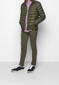 Green puffer jacket, plaid red and white button-up shirt, olive pants, and black sneakers with white accents. Textured materials and fitted design.