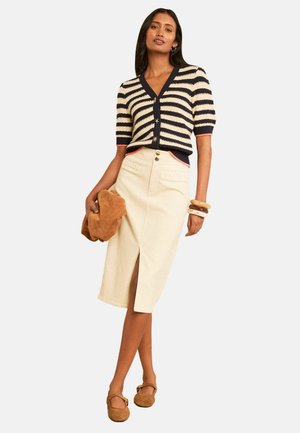 Frau trägt einen gestreiften kurzärmeligen Cardigan, cremefarbenen Midirock mit vorderem Schlitz, braune Schuhe, hält eine braune Clutch und trägt Armbänder.