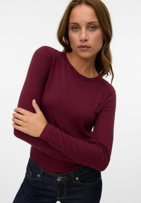 Femme aux cheveux châtain clair portant un haut moulant à manches longues en ribbed bordeaux et un jean bleu foncé, posant devant un fond blanc uni.