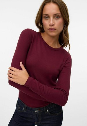 Vrouw met lichtbruin haar, gekleed in een aansluitend, lange mouwen ribbelachtige bordeaux top en donkerblauwe jeans, poserend voor een effen witte achtergrond.