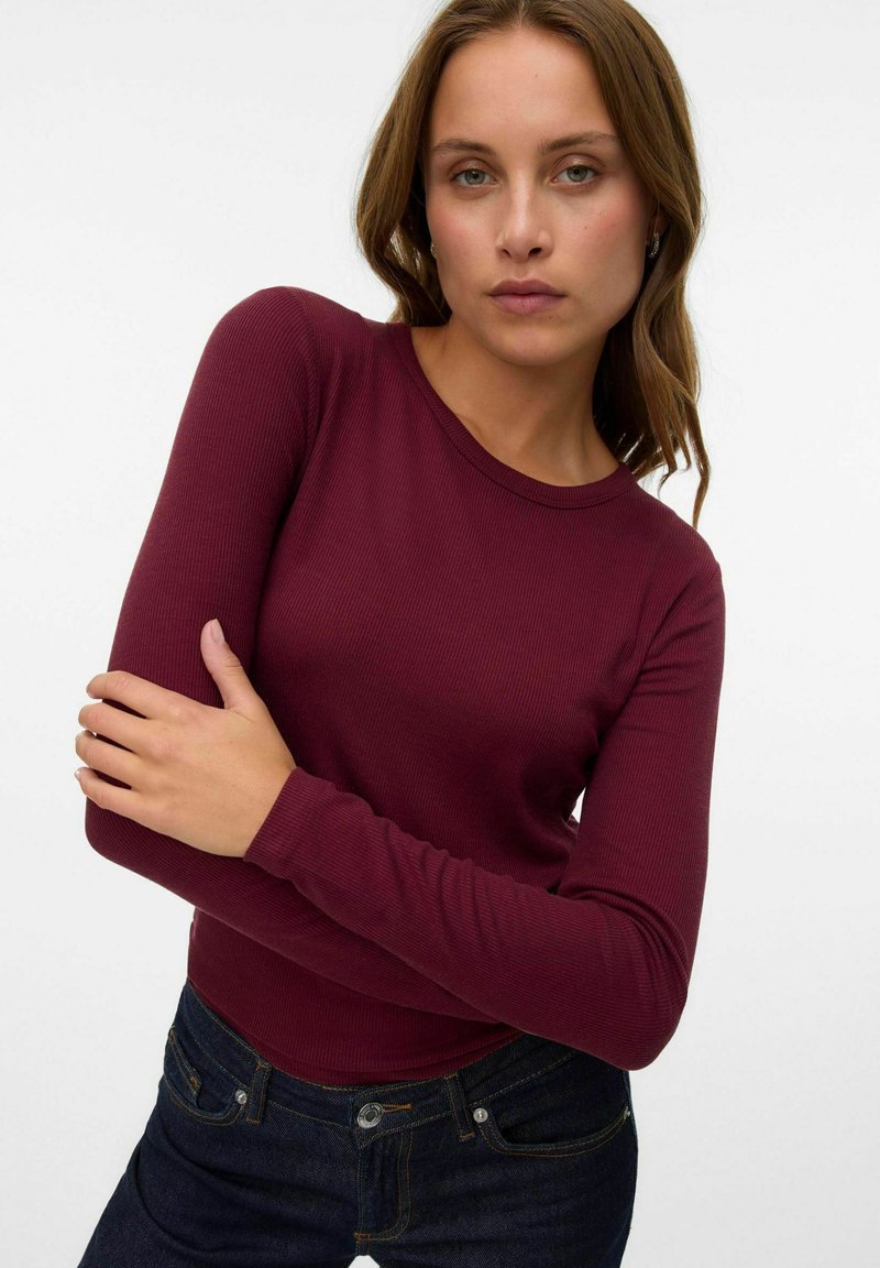 Femme aux cheveux châtain clair portant un haut moulant à manches longues en ribbed bordeaux et un jean bleu foncé, posant devant un fond blanc uni.