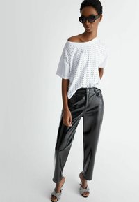 Top bianco a maniche corte con borchiette argentate, abbinato a pantaloni finti in pelle neri lucidi. La modella indossa occhiali da sole e scarpe aperte decorate.
