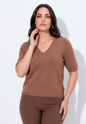 Vrouw met donker haar die een bruine korte mouw V-hals top en bijpassende broek draagt, poseert met één hand bij haar hals en een neutrale uitdrukking.
