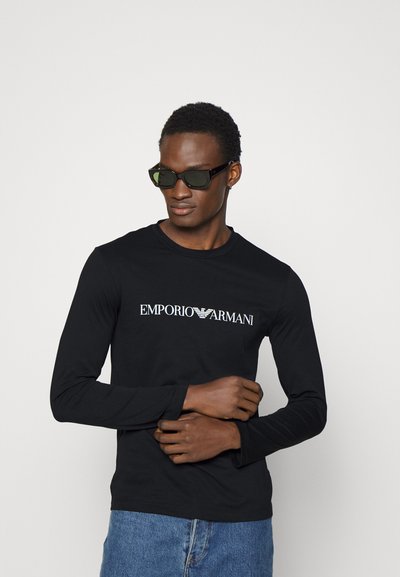 Juodas ilgomis rankovėmis marškinėliai su balta "EMPORIO ARMANI" užrašu, derinami su stačiakampiais tamsiais akiniais nuo saulės ir mėlynais džinsais.